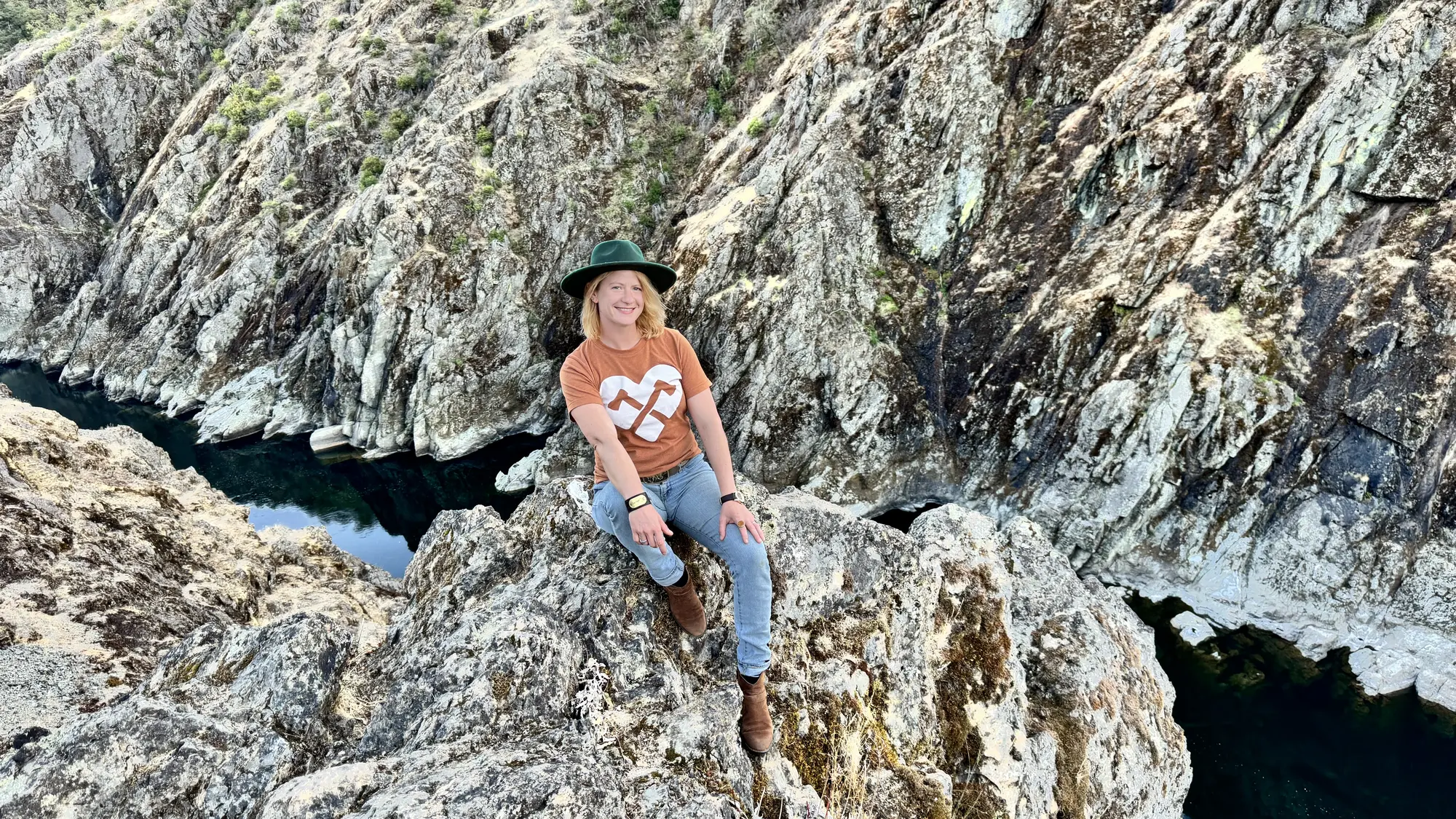 Sasha in a green hat and rust shirt, sitting on a rock high above a river canyon near her home in Southern Oregon